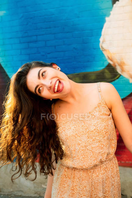 Playful woman in dress having fun and tilting head on side while standing by urban brick wall with graffiti — Stock Photo