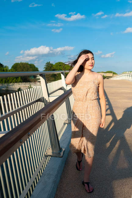Mujer relajada en vestido de sol paseando por el puente en el soleado día ventoso - foto de stock