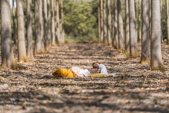 Side view of happy pregnant woman and little girl hugging and resting on ground in autumn forest glade — Stock Photo