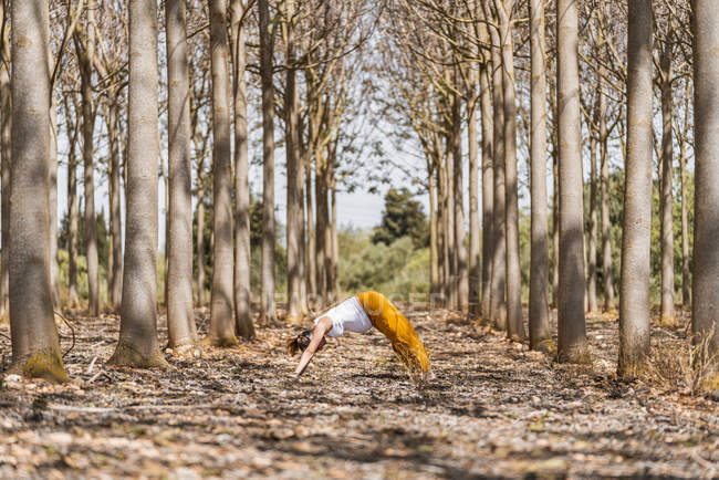 Vista lateral de la mujer embarazada en pantalones amarillos de pie en pose descendente mientras practica yoga en el suelo en el bosque - foto de stock