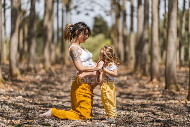 Side view of cheerful barefoot pregnant woman and little girl holding hands in autumn forest glade — Stock Photo