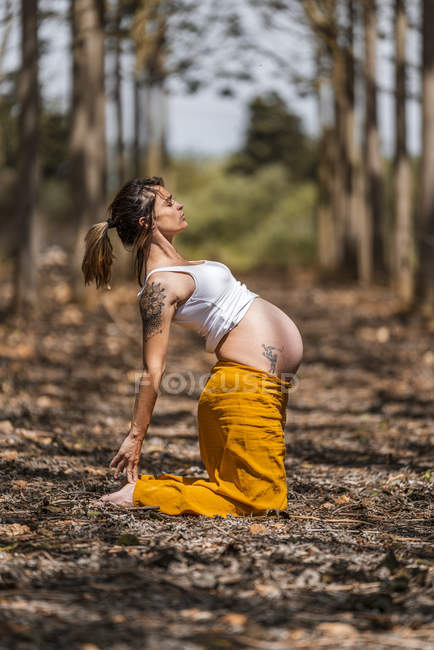 Vista lateral de la hembra embarazada con los ojos cerrados en camisa blanca y pantalones amarillos de pie sobre las rodillas mientras yoga en glade en el bosque - foto de stock