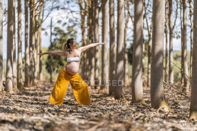 Взрослая беременная женщина в белой рубашке и желтых брюках стоит с протянутыми руками и практикует йогу среди деревьев — стоковое фото