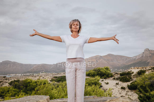 Woman clasping hands over head and closing eyes while standing on rock in nature and meditating — Stock Photo