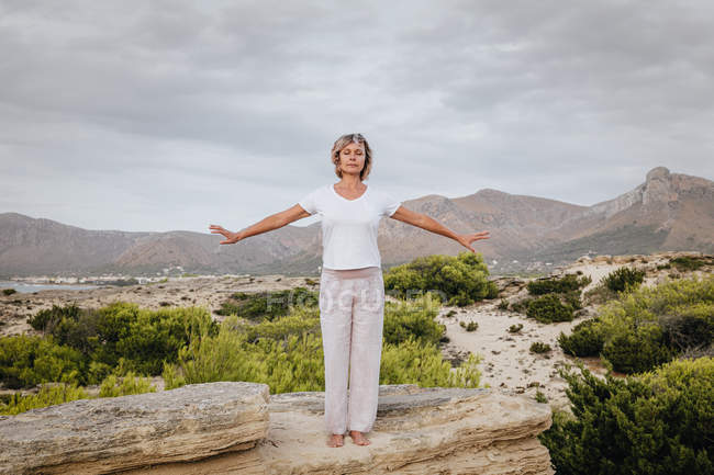 Mulher apertando as mãos sobre a cabeça e fechando os olhos enquanto está de pé sobre a rocha na natureza e meditando — Fotografia de Stock