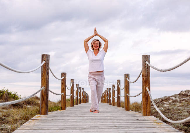 Woman smiling and clasping hands over head while standing on wooden path and practicing Tai Chi against overcast sky — Stock Photo