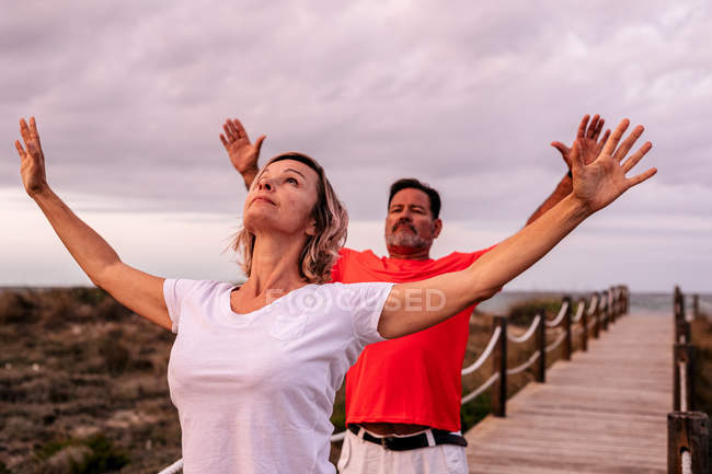 Adult man and woman stretching out arms and breathing while standing on wooden path against overcast sky — Stock Photo