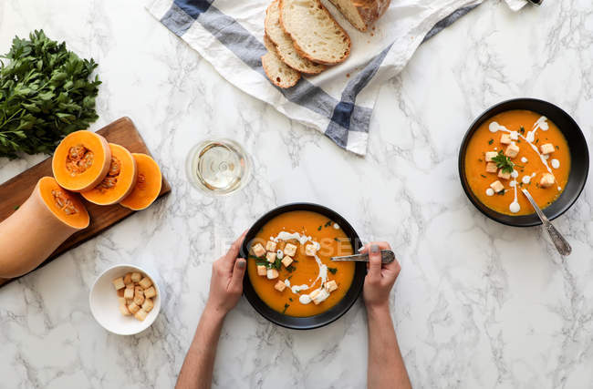 Da sopra mani di persona irriconoscibile che tiene la boccia di minestra di zucca arancione contro tavolo di marmo con verdure — Foto stock