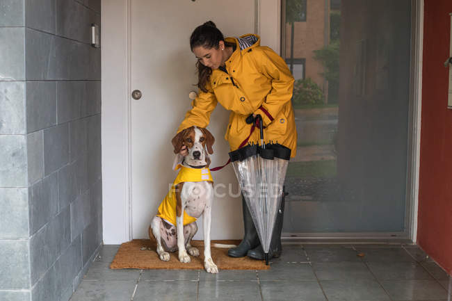 Obedient English Pointer in yellow cloak sitting at door with careful woman in yellow raincoat in entrance of house — Stock Photo