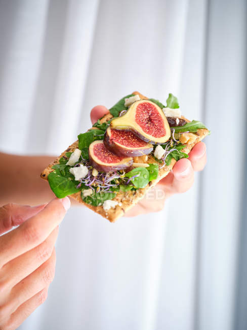 Person holding homemade open sandwich with slices of fig and cheese on rye bread with rocket salad — Stock Photo