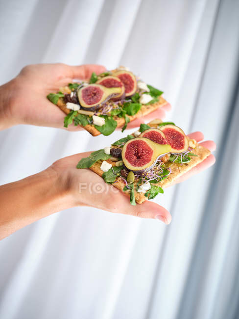 Person holding homemade open sandwiches with slices of fig and cheese on rye bread with rocket salad — Stock Photo