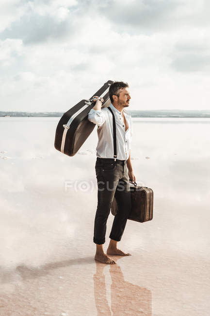 Vista lateral del hombre en camisa blanca y tirantes llevando la guitarra y el maletín mientras está de pie descalzo en el agua por la orilla - foto de stock