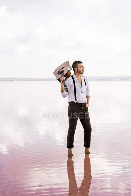 Passionate man in white shirt and suspenders holding guitar while standing barefoot in water by shore on cloudy weather — Stock Photo