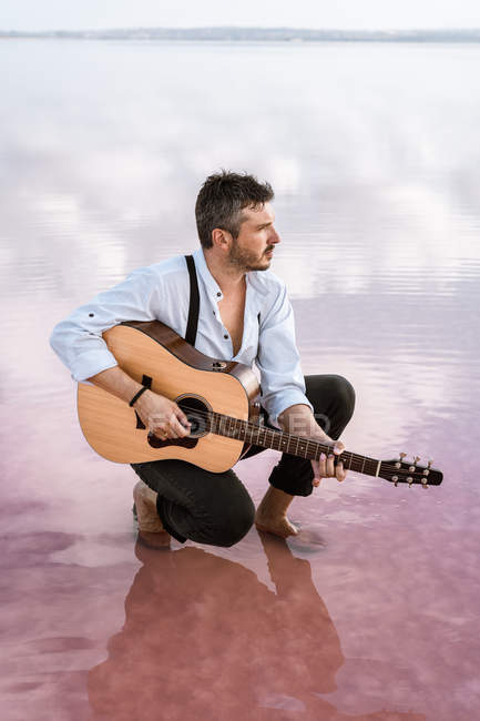 Uomo malinconico in camicia bianca e bretelle che suonano la chitarra acustica e distolgono lo sguardo circondato da acqua di mare liscia — Foto stock