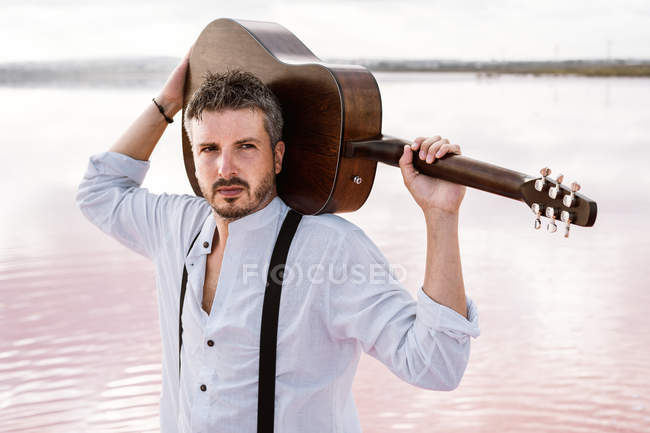 Man in white shirt holding acoustic guitar while standing on shore on cloudy daytime while looking away — Stock Photo