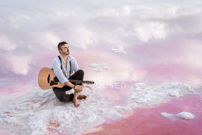 Сміливий чоловік з акустичною гітарою сидить на пляжі, дивлячись далеко в оточенні гладкого моря, що відображає величний хмарний пейзаж — стокове фото