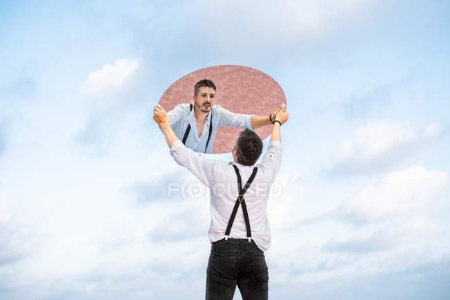 Відбиття низького кута стомленого чоловіка в сорочці і підвісках, що стоять над блакитним небом і піднімає овальне дзеркало — стокове фото