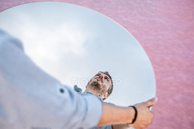 From above reflection of pensive man in shirt and suspenders standing over blue sky and lifting oval mirror — Stock Photo