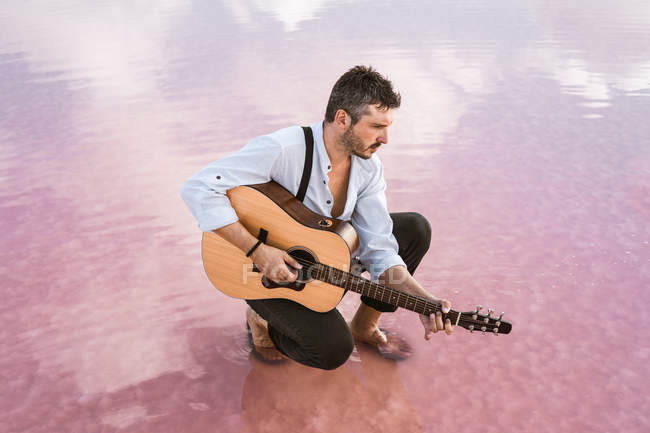 Uomo malinconico che suona la chitarra acustica seduto sulla spiaggia circondato da un mare liscio che riflette maestoso paesaggio nuvoloso — Foto stock