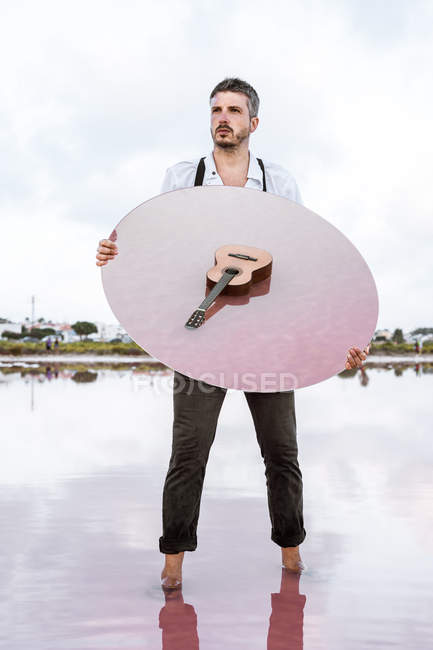 Homem segurando espelho oval com reflexo de guitarra em água rosa — Fotografia de Stock
