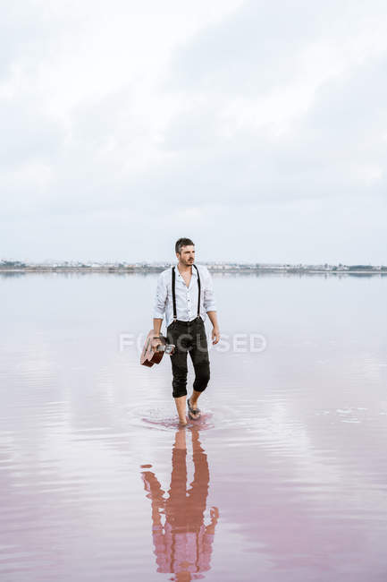 Hombre de camisa blanca y tirantes sosteniendo la guitarra mientras está de pie descalzo en el agua por la orilla en el día nublado - foto de stock