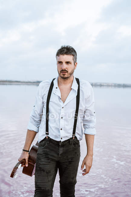 Чоловік у білій сорочці і підтяжки тримають гітару, стоячи у воді на березі в похмурий день — стокове фото