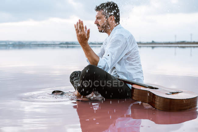 Man in white shirt and suspenders throwing water while sitting on beach with pink water near acoustic guitar — Stock Photo