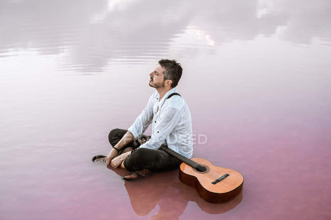 Hombre melancólico con guitarra acústica sentado en la playa mirando hacia otro lado rodeado de mar suave que refleja majestuoso paisaje nublado - foto de stock
