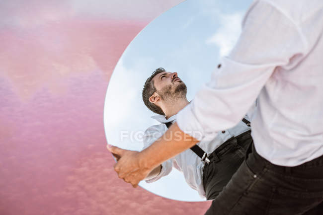 De cima a reflexão do homem pensativo na camisa e suspensórios que estão sobre o céu azul e levantam o espelho oval — Fotografia de Stock