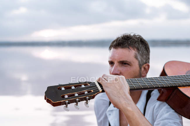 Hombre de camisa blanca y tirantes llevando guitarra acústica y sentado en la playa por el agua - foto de stock