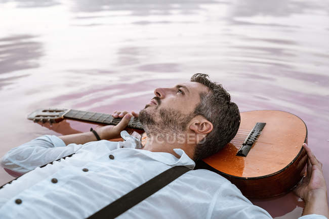Сверху человек в белой рубашке и подтяжках, лежащих на плавающей акустической гитаре в море на песчаном берегу — стоковое фото