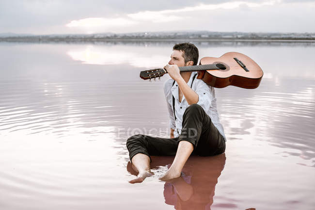 Wistful man in white shirt and suspenders carrying acoustic guitar and sitting on beach with pink water — Stock Photo