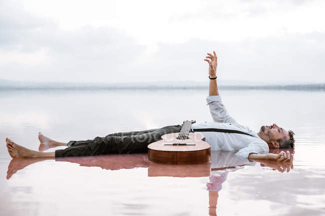 Mann mit Gitarre auf Rücken liegend, wegschauend und ausgestreckte Hand zum Himmel im Meer auf Sandbank — Stockfoto