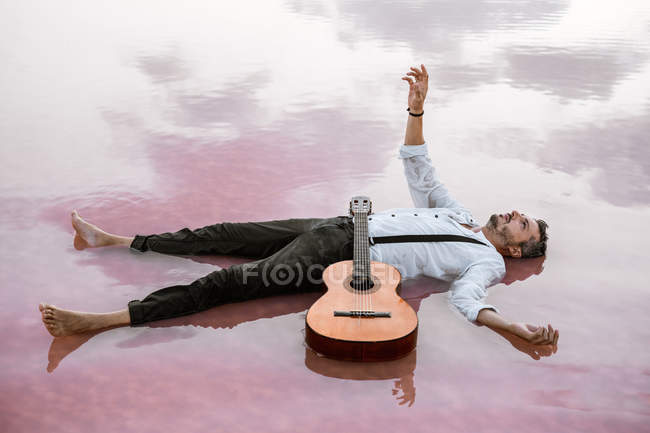 Человек с гитарой лежит на спине, глядя в сторону и протягивая руку к небу в море на песчаном берегу — стоковое фото
