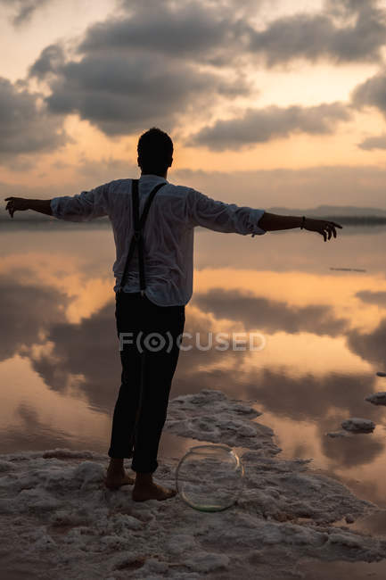Ззаду чоловік у мокрому одязі дивиться і стоїть з витягнутими руками з порожнім рибним баком на узбережжі в хмарному заході сонця — стокове фото