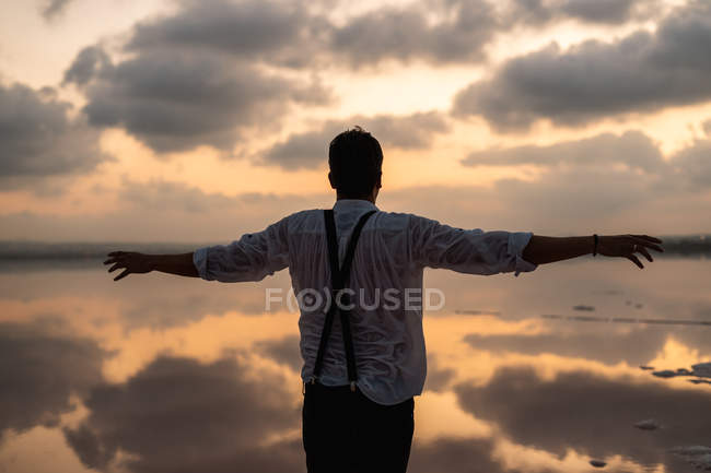 Von hinten Mann in nassen Kleidern starrt und steht mit ausgestreckten Armen an der Küste im wolkenverhangenen Sonnenuntergang — Stockfoto