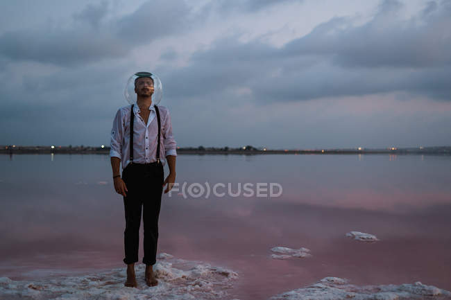 Страшный человек с закрытыми глазами в мокрой рубашке с пустым аквариумом на голове стоя и созерцая неподвижное море в сумерках — стоковое фото