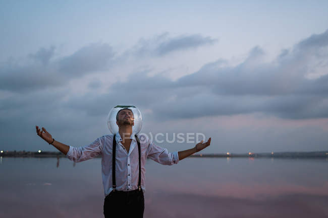 Pensive man with closed eyes in wet shirt with empty aquarium on head standing with outstretching arms by seaside in twilight — Stock Photo