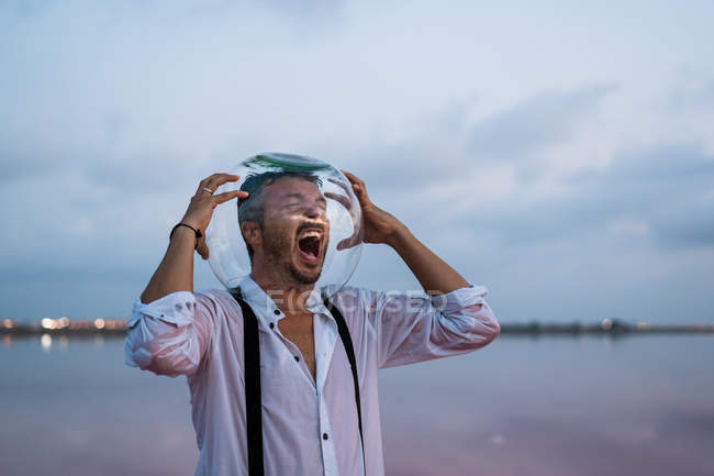 Distressed man in agony in wet shirt with empty aquarium on head standing screaming by still sea in twilight — Stock Photo