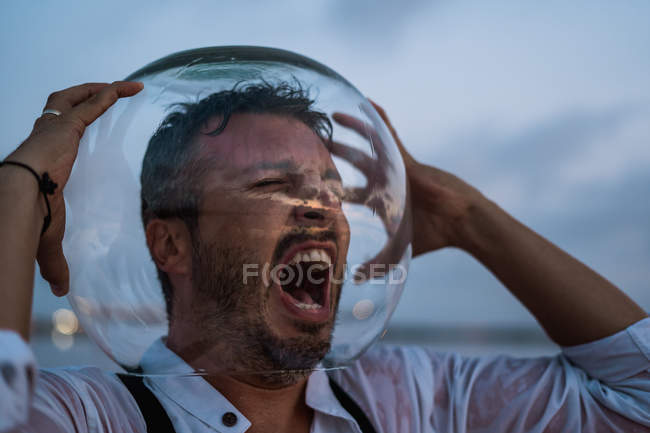 Distressed man in agony in wet shirt with empty aquarium on head standing screaming by still sea in twilight — Stock Photo