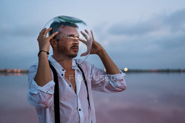 Pensive man in wet shirt with empty aquarium on head standing and contemplating by still sea in twilight — Stock Photo