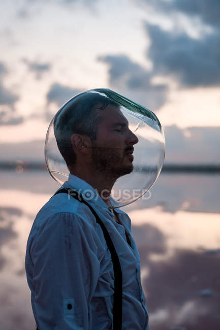 Pensive man with closed eyes in wet shirt with empty aquarium on head standing and contemplating by still sea in twilight — Stock Photo
