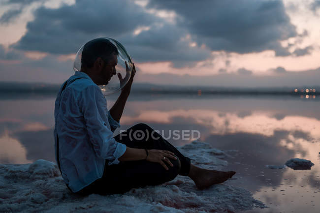 Nachdenklicher Mann im nassen Hemd holt leeres Aquarium hervor, während er still am Meer in der Dämmerung sitzt — Stockfoto