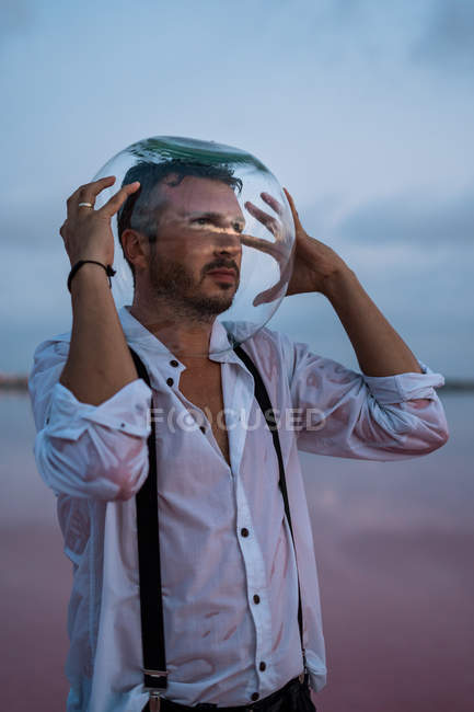 Людина в мокрій сорочці з порожнім акваріумом на голові стоїть і розмірковує над морем в сутінках — стокове фото