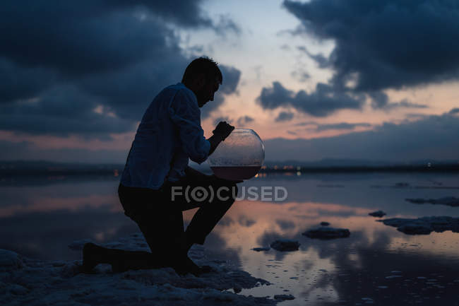 Vista lateral del hombre arrodillado y llenando el acuario vacío con agua del océano en la playa en el crepúsculo - foto de stock