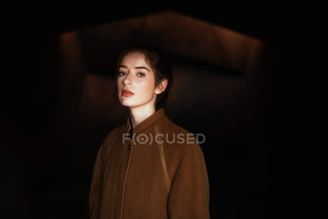 Donna pensierosa in cappotto marrone guardando in macchina fotografica contro sfondo nero — Foto stock