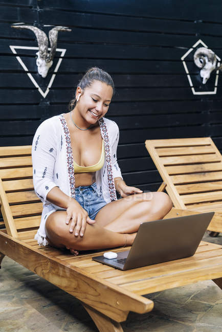 Fröhliche Frau in legerer Kleidung blättert am Laptop, während sie mit gekreuzten Beinen auf einem hölzernen Liegestuhl auf der tropischen Terrasse sitzt — Stockfoto