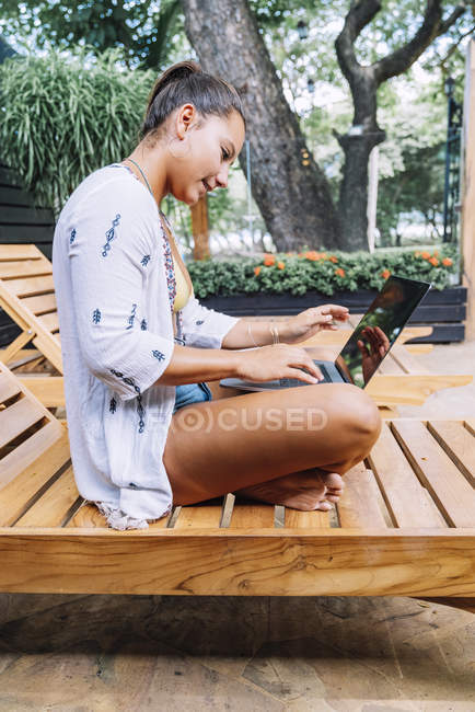 Вид збоку жінки в повсякденному одязі переглядає ноутбук, сидячи з схрещеними ногами на дерев'яному стільці на тропічній терасі — стокове фото