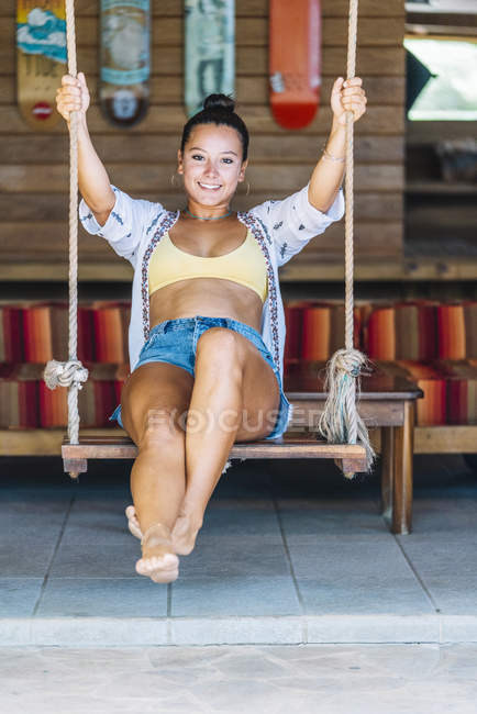 Tanned happy woman in casual wear sitting on swings and looking at camera over wooden wall in Costa Rica — Stock Photo