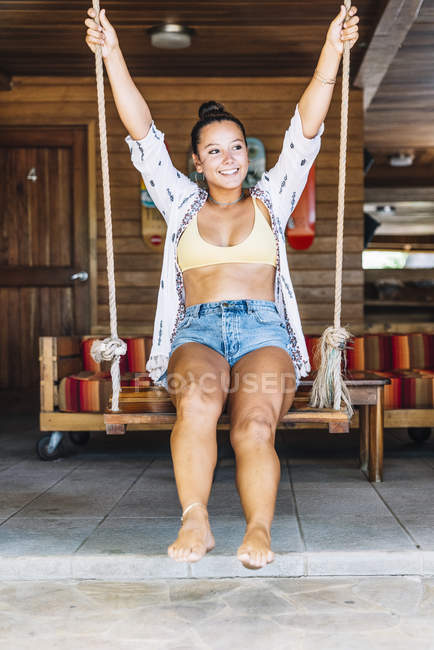 Mulher feliz bronzeada no desgaste casual que se senta em balanços e olha afastado sobre a parede de madeira em Costa Rica — Fotografia de Stock